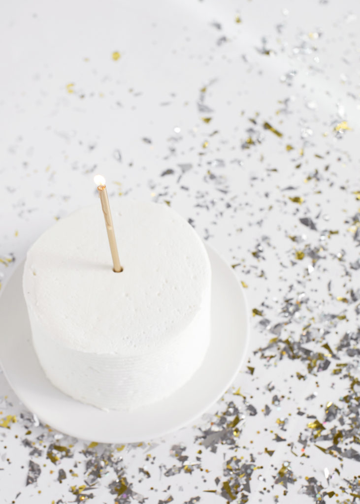 White cake with a single lit candle on a white plate on a table covered in confetti